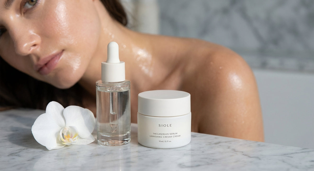 A luminous close-up of dewy, radiant skin alongside a glass serum bottle and cream jar on a cool marble surface, lit with soft natural light.
